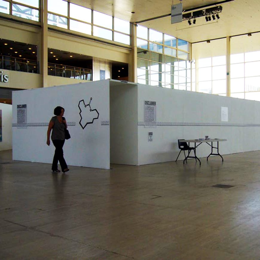 Photograph of visitor approaching white exhibition structure with Disclaimer sign and table by doorway