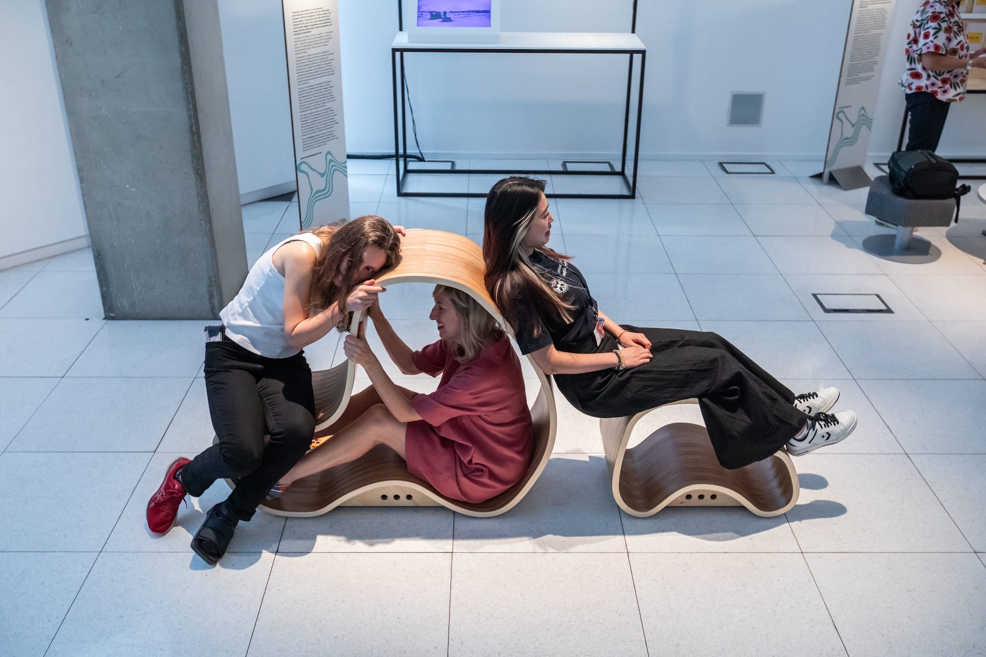 Three adult females lying, leaning and sitting inside two organically shaped wooden forms in a gallery