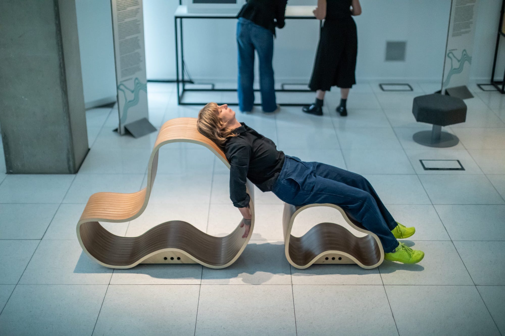 Adult female lying across two organically shaped wooden forms in a gallery